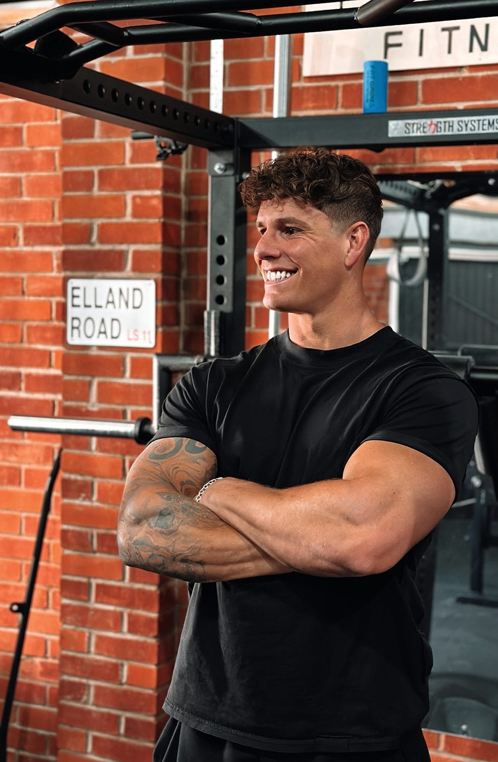 Luke Hayes laughing with his arms folded in a brick-walled gym