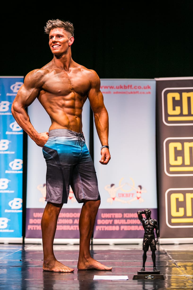 Luke on stage at a UKBFF physique competition, with the trophy at his feet